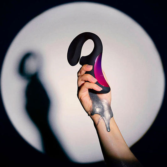 Mano sosteniendo el vibrador Enigma™ de LELO con diseño elegante en negro y detalles en color púrpura, capturado frente a un fondo circular iluminado.